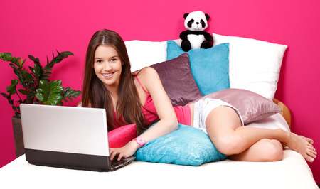 Teenage girl lying on bed with pillows, stuffed panda, typing on laptop in a pink bedroom